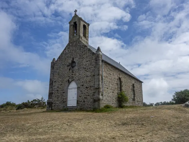 Église en pierre avec un clocher et une porte blanche, sous un ciel bleu et nuageux