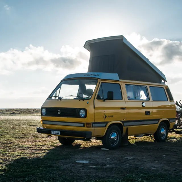 Ein gelber Wohnwagen mit aufgestellter Dachluke ist am Strand geparkt, daneben steht ein Fahrrad