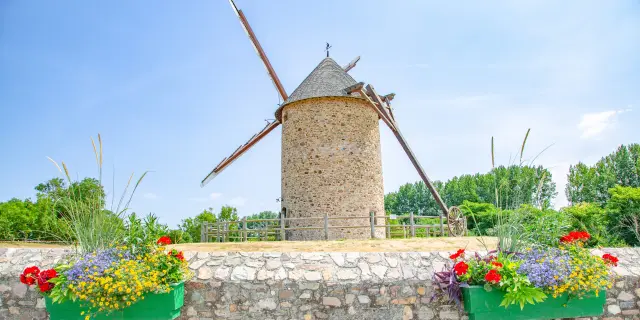 Un moulin à vent en pierre avec des ailes en bois, entouré de fleurs colorées dans des jardinières vertes.