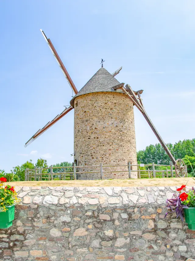 Un moulin à vent en pierre avec des ailes en bois, entouré de fleurs colorées dans des jardinières vertes.
