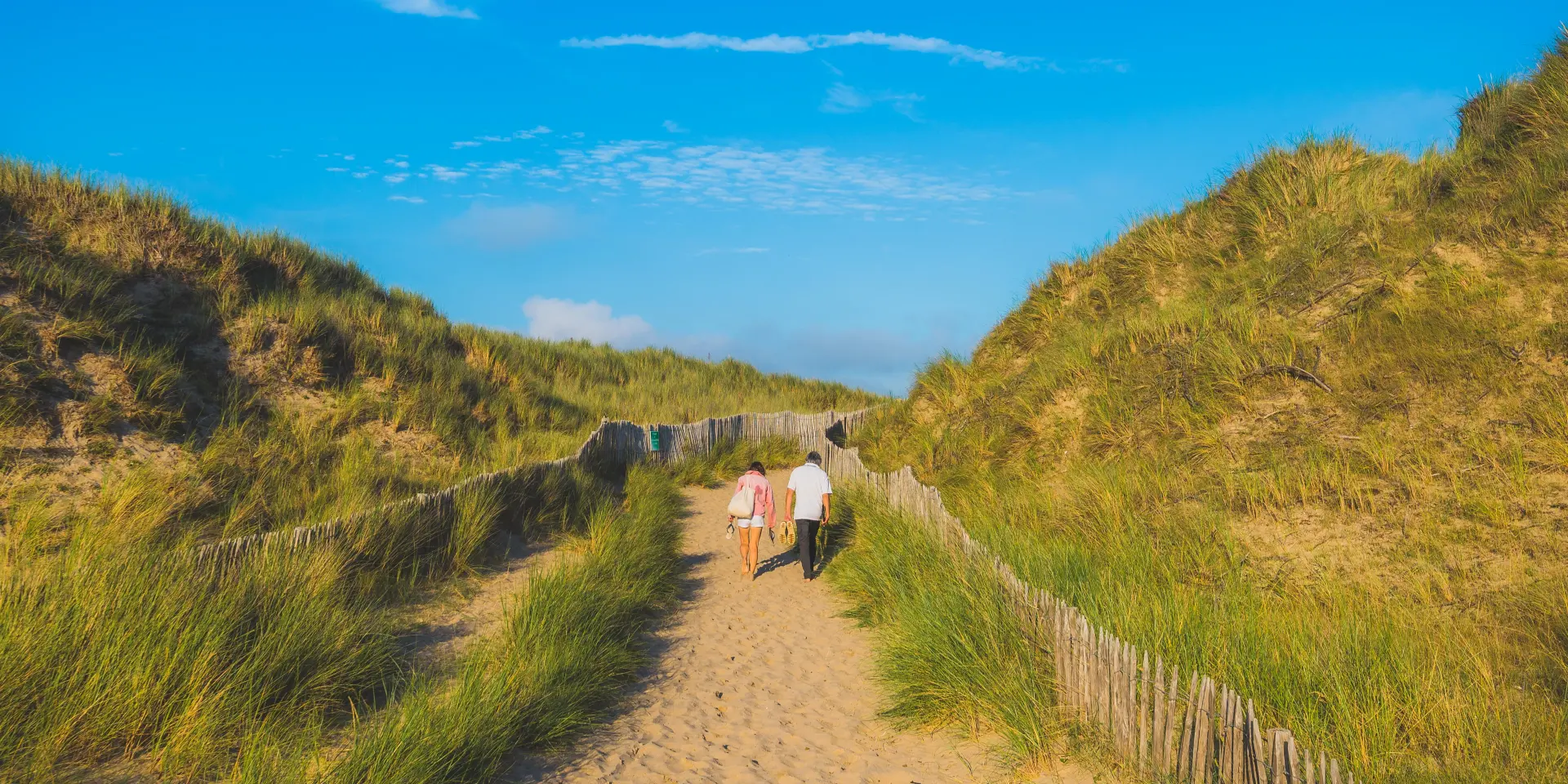 Zwei Erwachsene und ein Kind gehen auf einem Sandweg zwischen grasbewachsenen Dünen