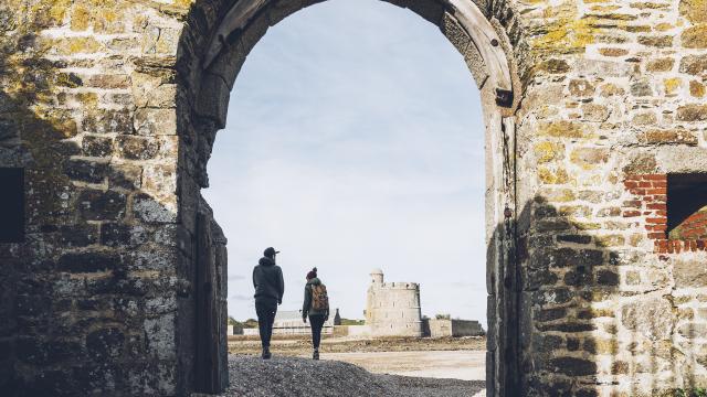 Two people walking through a ruined archway towards a distant tower