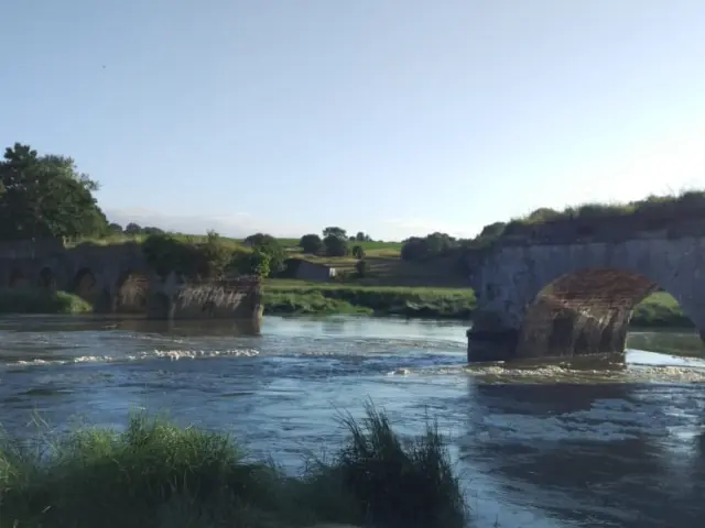 Eine Steinbrücke, die einen Fluss überspannt, mit Bäumen und Feldern im Hintergrund