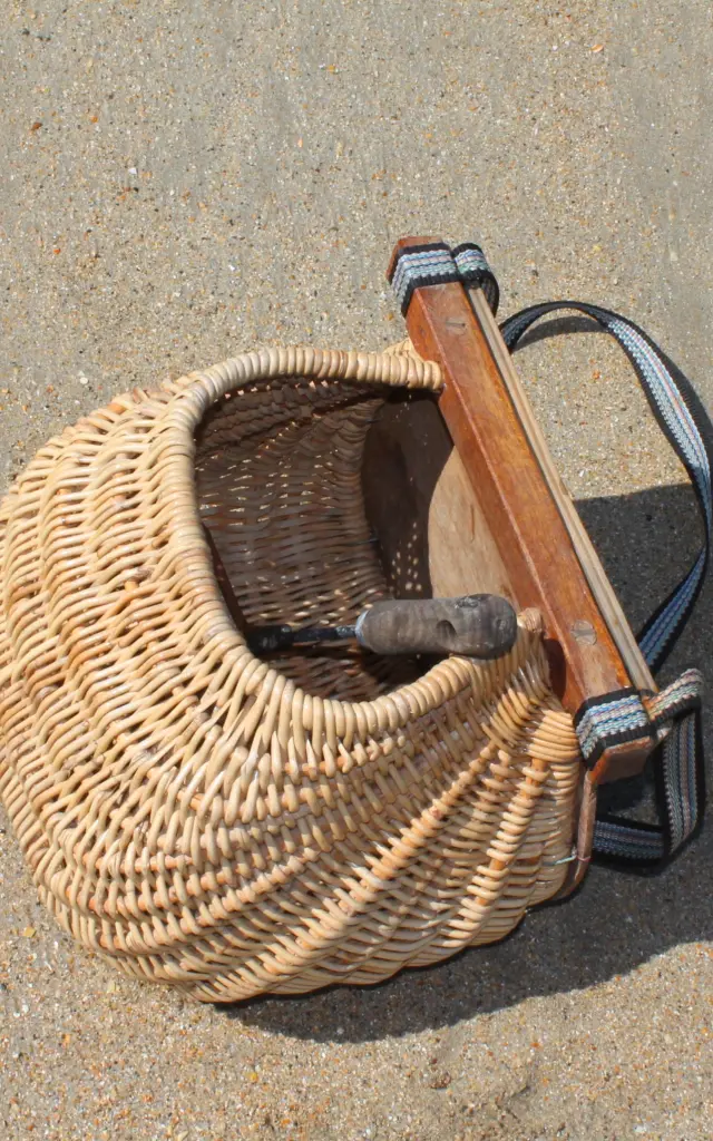 Un panier en osier de pêche à pied avec une poignée en bois et une sangle noire posé sur le sable
