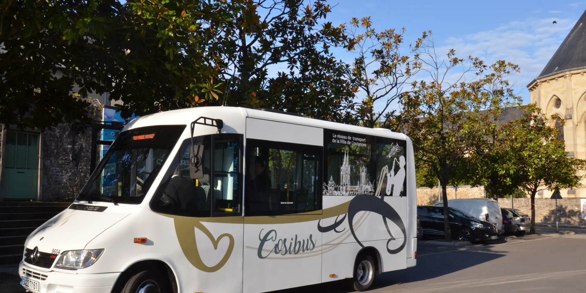 Ein weißer Stadtbus mit schwarzen und goldenen Mustern fährt eine von Bäumen gesäumte Straße entlang.