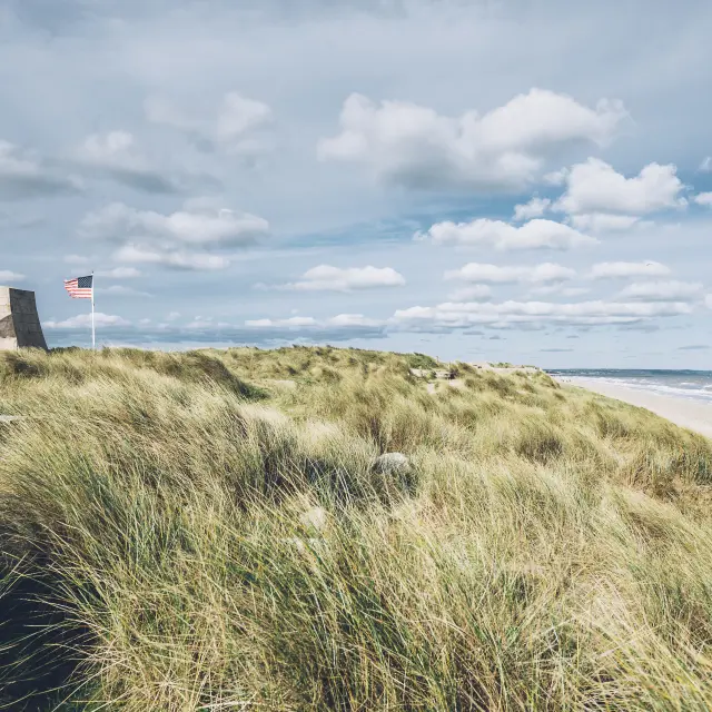 Sand dunes with two flags, one French and one American, fluttering in the wind near the ocean