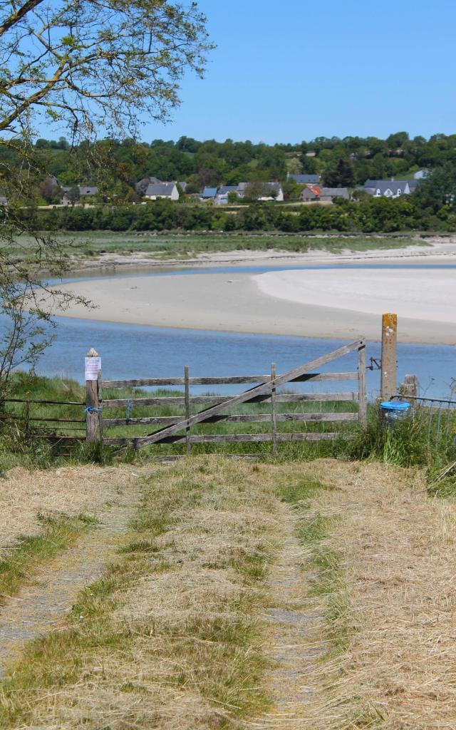 A sandy beach with a village in the background and a path leading to a wooden fence