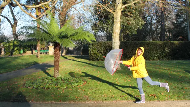 Une femme en ciré et bottes grises marche dans un parc avec un parapluie transparent
