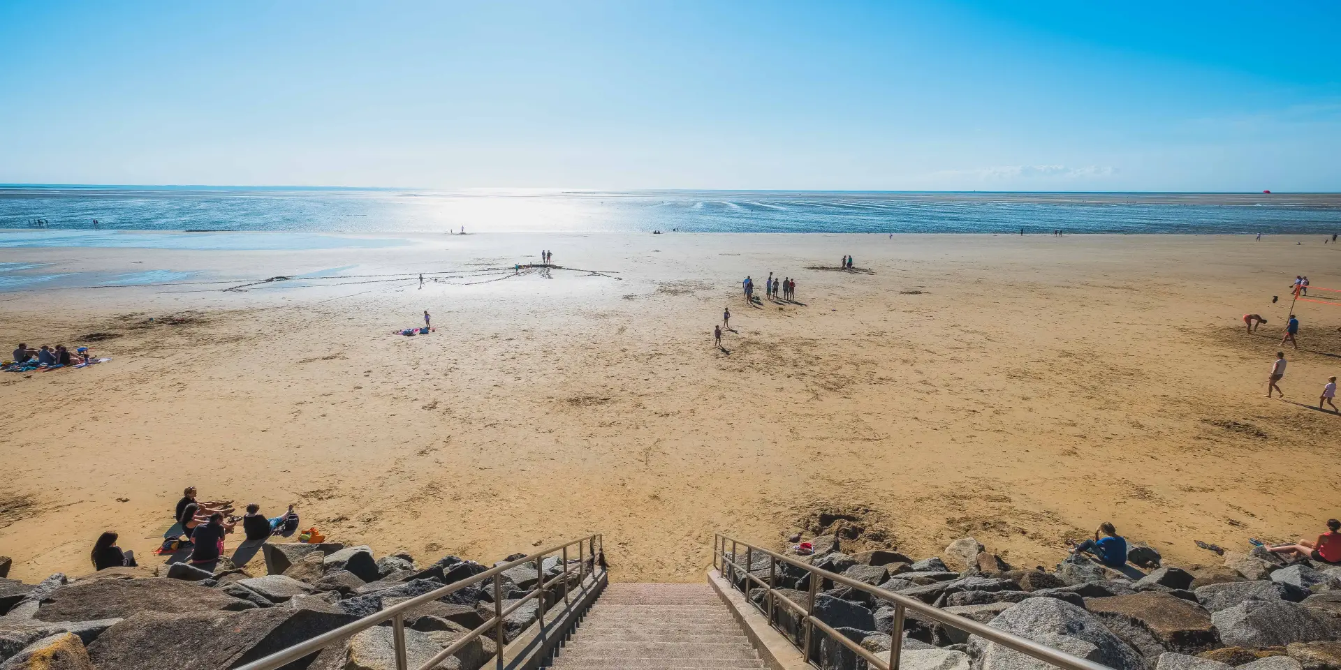 Stairs leading to a sandy beach with people relaxing and playing near the water