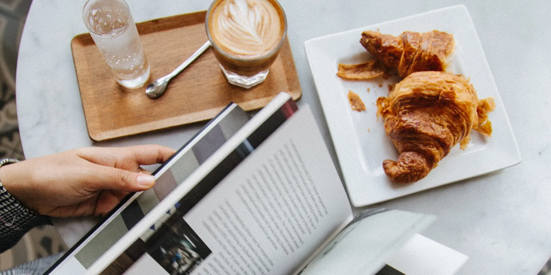 Personne lisant un magazine à une table avec un café et des croissants