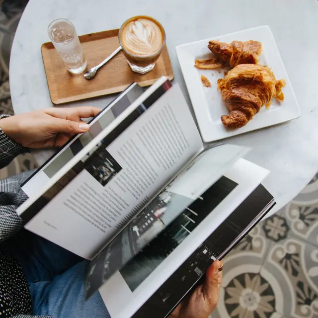 Personne lisant un magazine à une table avec un café et des croissants