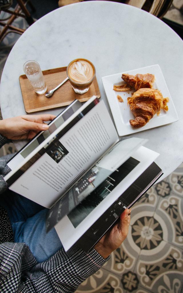 Personne lisant un magazine à une table avec un café et des croissants