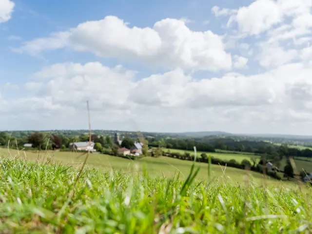 Champ vert avec des maisons et des collines à l'horizon sous un ciel bleu avec des nuages blancs