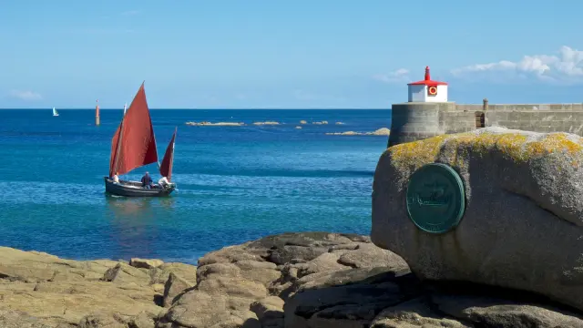 A red sailboat sails near a rocky coastline with a lighthouse