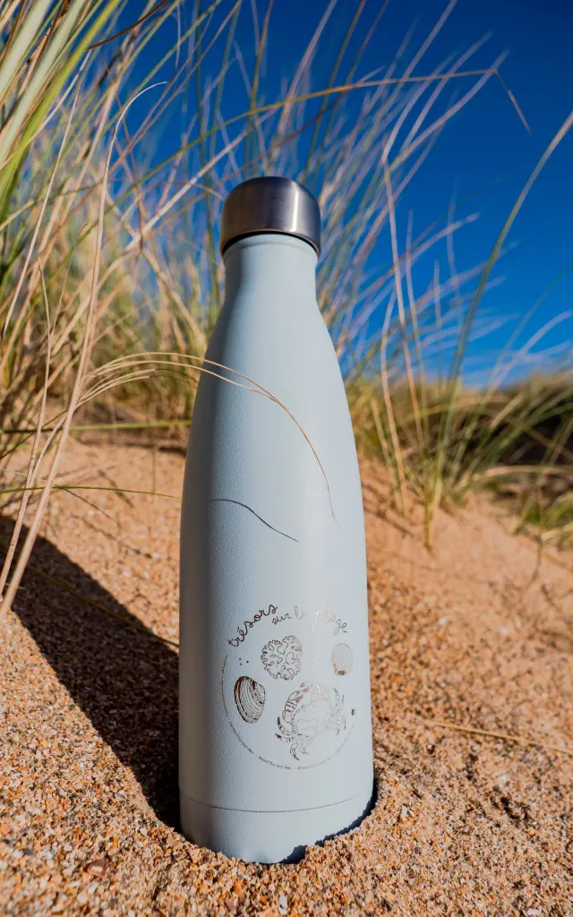Une gourde en acier inoxydable sur le sable avec des herbes autour