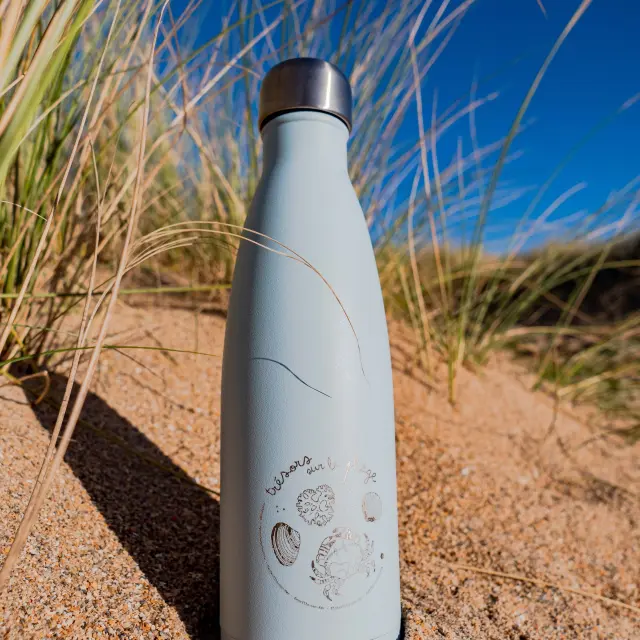Une gourde en acier inoxydable sur le sable avec des herbes autour