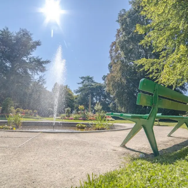 Un banc vert dans un jardin avec une fontaine au centre au jardin des plantes de Coutances