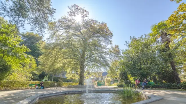 Un jardin avec une fontaine au centre, des arbres et des personnes assises sur des bancs : jardin des plantes de Coutances