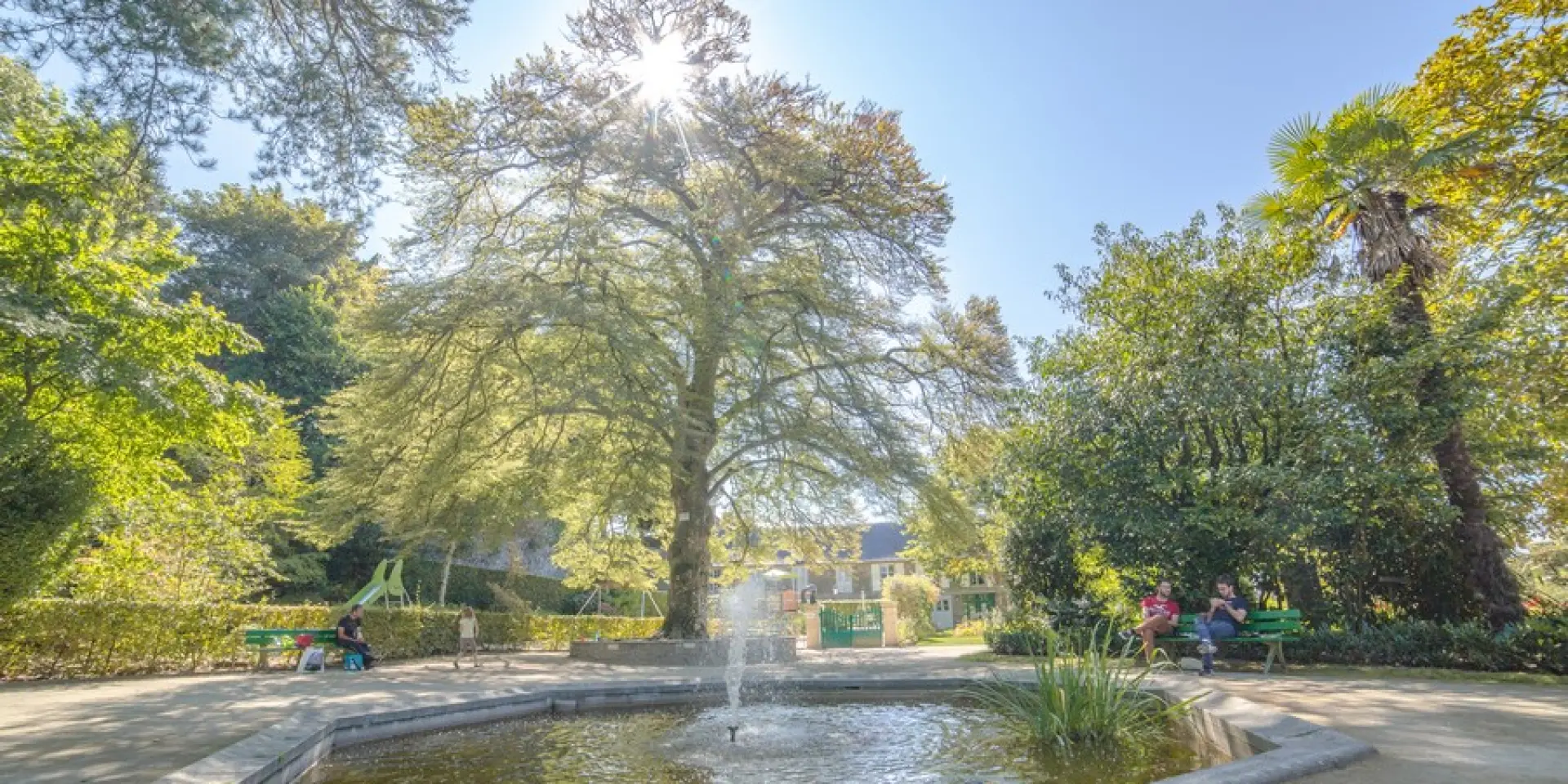 Un jardin avec une fontaine au centre, des arbres et des personnes assises sur des bancs : jardin des plantes de Coutances