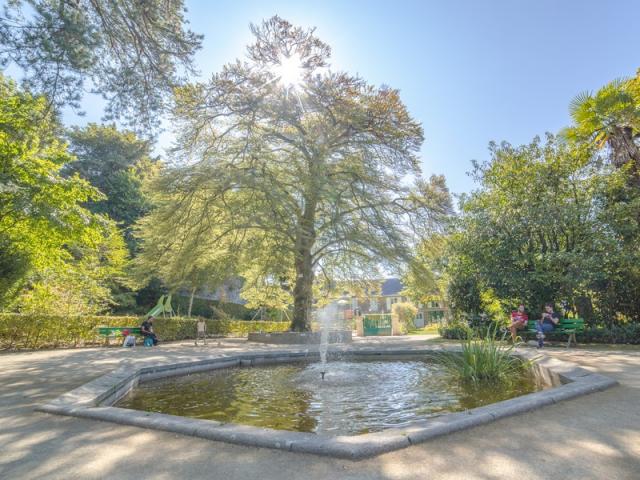 Un jardin avec une fontaine au centre, des arbres et des personnes assises sur des bancs : jardin des plantes de Coutances