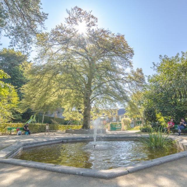 Un jardin avec une fontaine au centre, des arbres et des personnes assises sur des bancs : jardin des plantes de Coutances