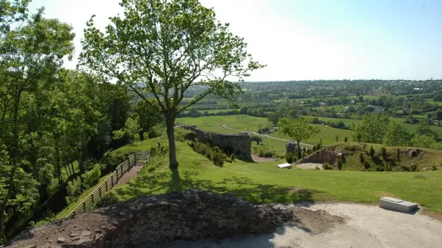Ein Feldweg führt über einen grünen Hügel mit alten Ruinen und Blick auf das Land