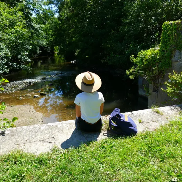 Une personne assise au bord d'une rivière, sur la voie verte à Bricqueville La Blouetteportant un chapeau et regardant l'eau