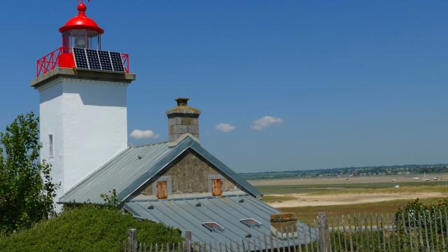 Weißer Leuchtturm mit einer roten Laterne und einem Solarpanel, angrenzend an ein graues Haus mit einem Metall-Dach
