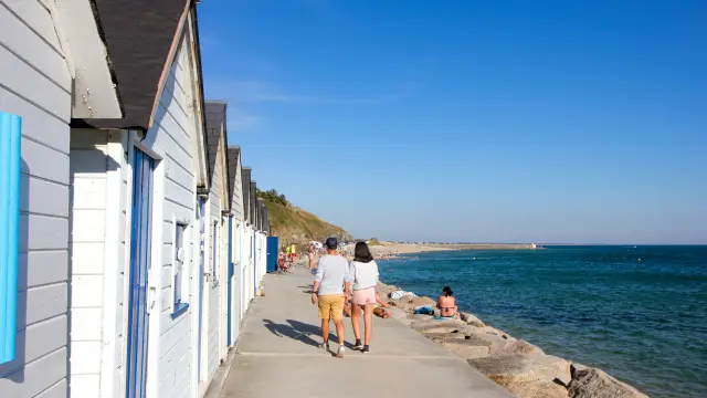 Two people walking along a seaside promenade with white beach huts
