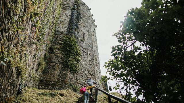 Deux personnes montent un escalier vers une tour en pierre couverte de végétation au château de Bricquebec