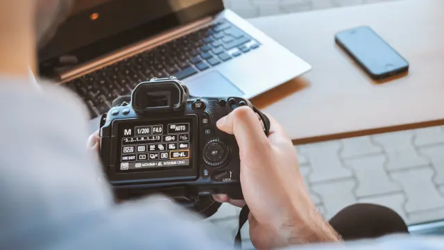 Une personne tenant un appareil photo devant un ordinateur portable et un smartphone sur une table.