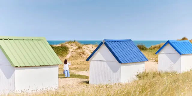 Three beach huts with differently colored roofs, green, blue, and white, on a grassy dune facing the sea.