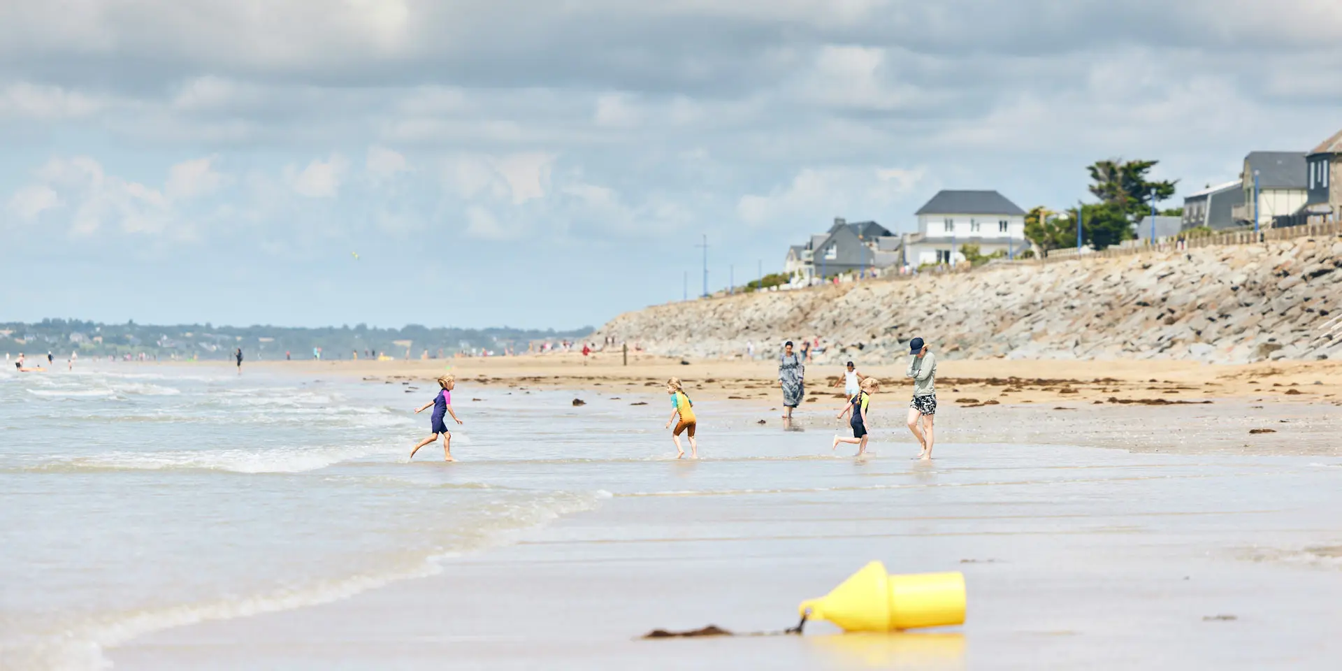 Plusieurs enfants courent sur une plage de sable avec des maisons en arrière-plan à Hauteville-sur-Mer