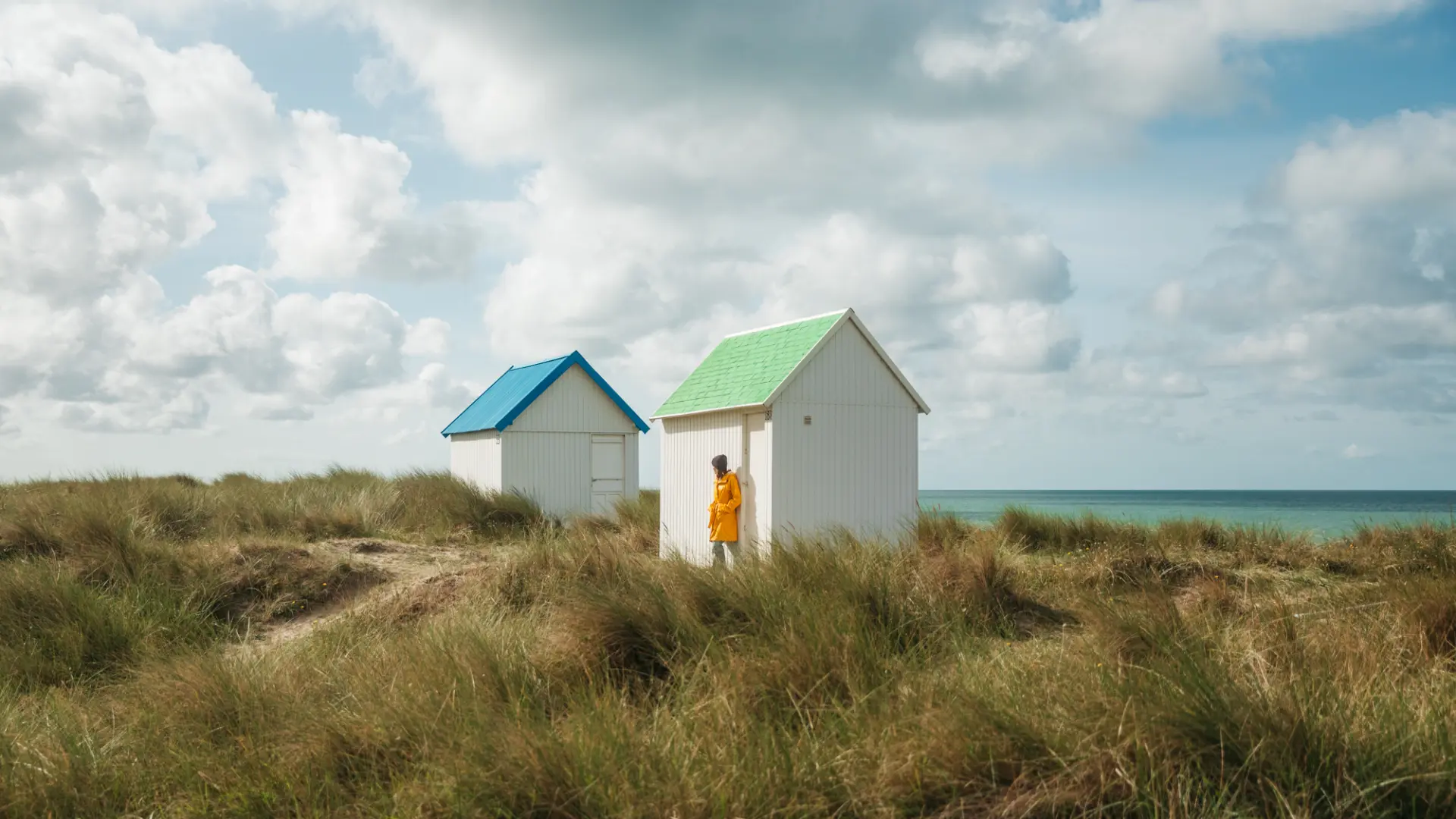 Two white beach huts with colorful roofs, one green and one blue, on a grassy dune near the sea.
