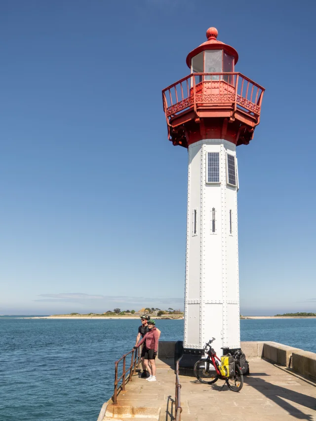 A white lighthouse with a red lantern on top, located by the water with people and bicycles nearby