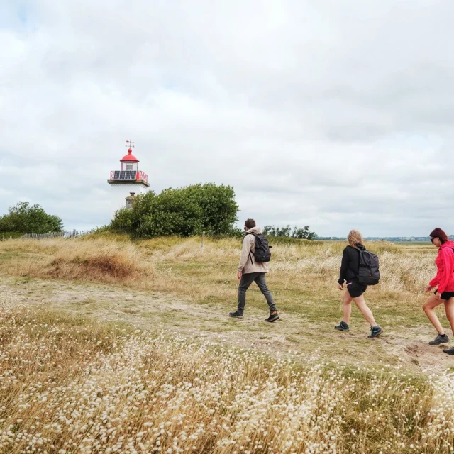 Trois personnes marchant vers un phare dans un champ herbeux de la pointe d'Agon