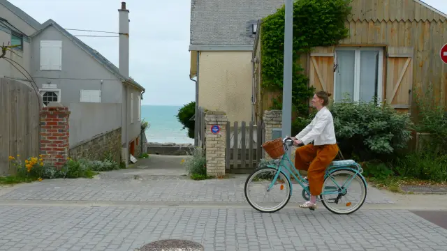 Une femme en blanc et orange roule à vélo sur une route pavée près de la mer à Agon-Coutainville