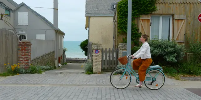 Une femme en blanc et orange roule à vélo sur une route pavée près de la mer à Agon-Coutainville