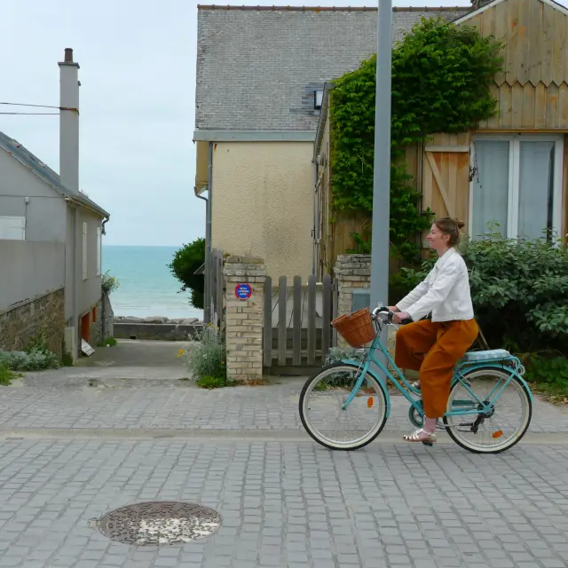 Une femme en blanc et orange roule à vélo sur une route pavée près de la mer à Agon-Coutainville