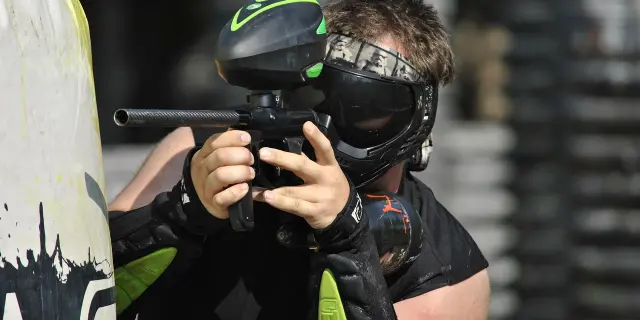 Un joueur de paintball vise avec son lanceur, portant un masque de protection et des gants. Paintball Saint Sauveur Villages